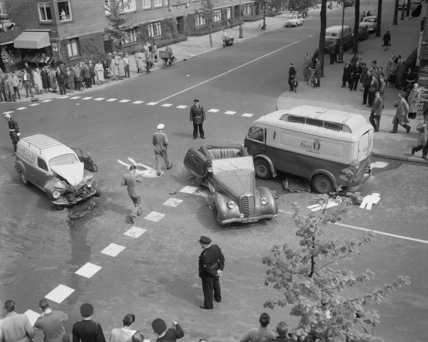 Schwarze und weiße Szene eines Autounfalls an einer Straßenkreuzung mit mehreren Fahrzeugen, darunter ein Van, und einer Gruppe von Menschen in der Nähe, mit Gebäuden, Bäumen, Pfählen und Texttafeln im Hintergrund.