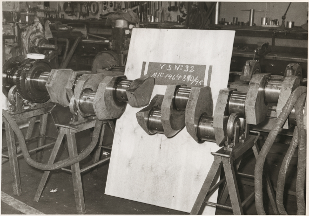 Ein Schwarz-Weiß-Foto einer Fabriksammlung mit Metallobjekten auf Ständern und einer Texttafel, mit verstreuten Werkzeugen und Objekten im Hintergrund.