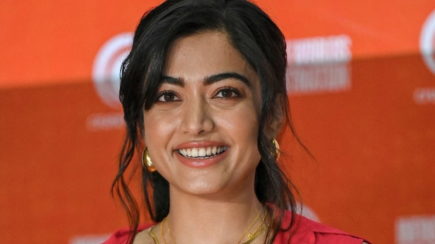 Frau in rotem Kleid mit elegantem Dutt und dezentem Make-up, die in die Kamera lächelt, vor einem gelben Banner mit der Aufschrift "Sonam Kapoors Schönste Looks vom Cannes Film Festival".