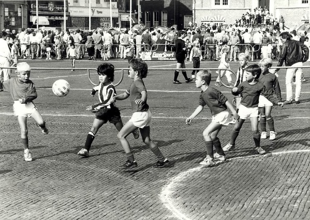 Schwarzes Bild von jungen Jungs, die auf der Straße Fußball spielen, umgeben von einer Zuschauermenge hinter einem Geländer, Gebäuden und Pfählen im Hintergrund.