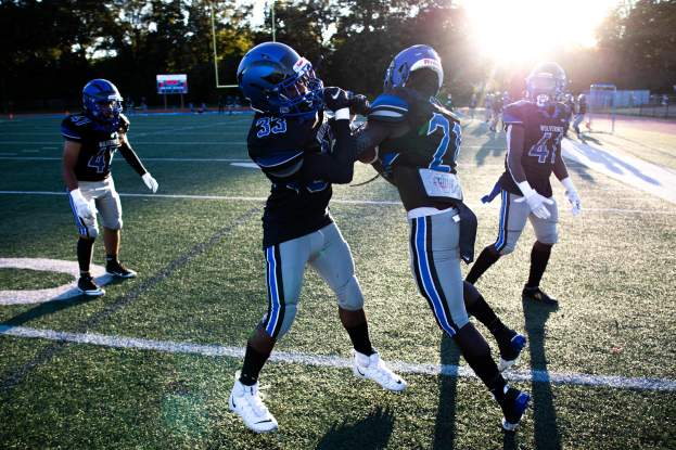 Eine Gruppe junger Männer, die Fußball auf einem von Bäumen umgebenen Feld unter einem klaren blauen Himmel spielen, tragen Helme und Schuhe.