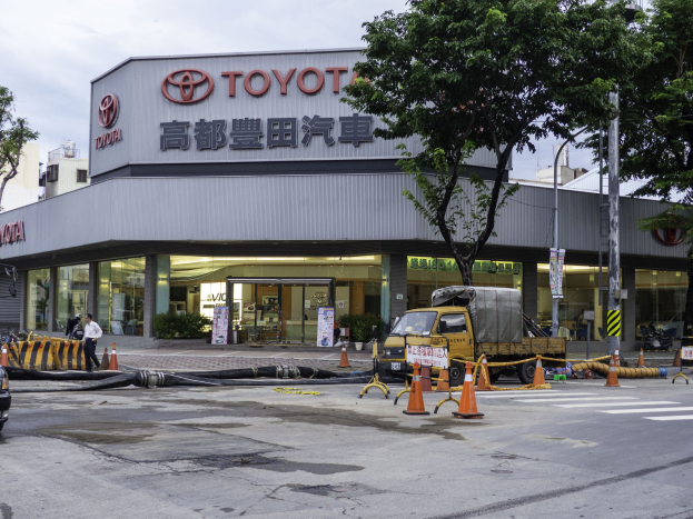 Außenansicht einer Toyota-Niederlassung in Tokio, Japan, mit Fahrzeugen, Verkehrskegeln, Rohren, Fußgängern, städtischer Infrastruktur und Gebäuden unter einem bewölkten Himmel.