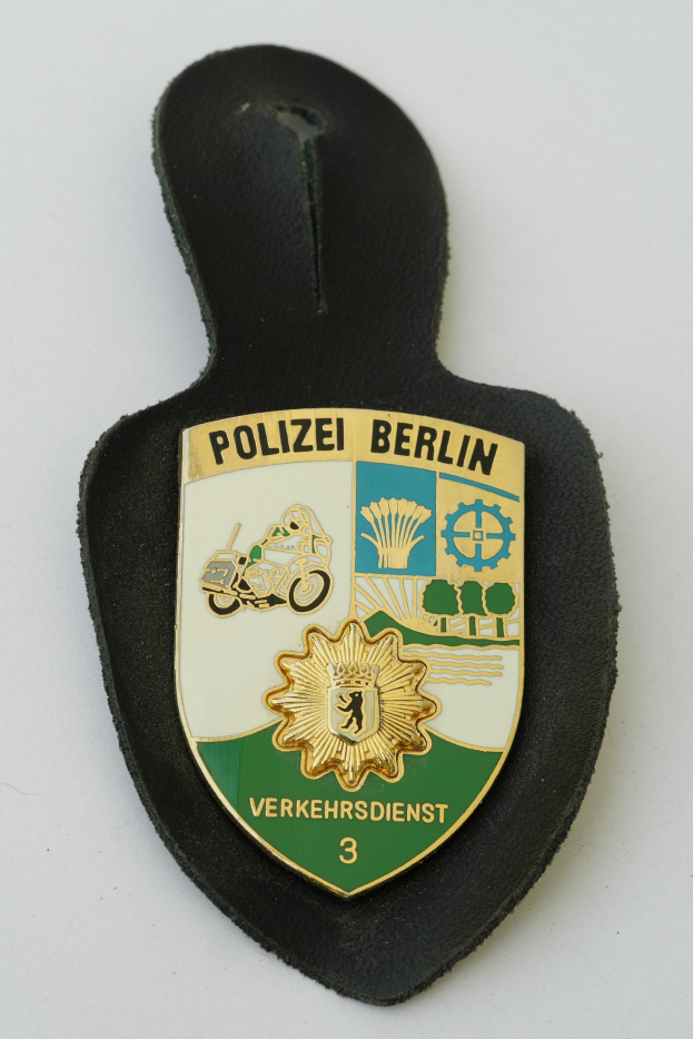 Ein schildförmiges Polizeibadges mit "Polizei Berlin" in schwarzer fettgeschriebener Schrift auf einem blauen Hintergrund mit einem weißen Stern und Rahmen, das auf einer Oberfläche liegt.