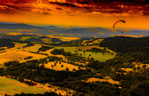 Zwei Paraglider über einem grünen Feld mit Bäumen, Häusern und Bergen im Hintergrund unter einem bewölkten Himmel.
