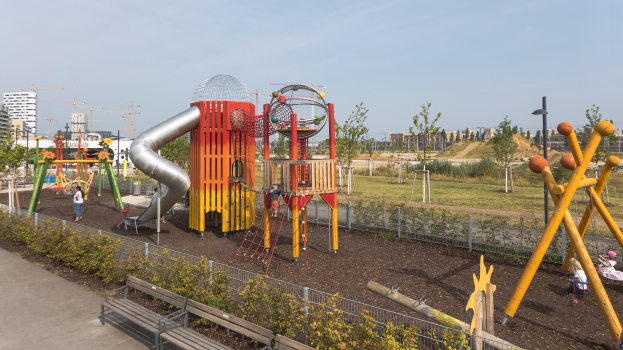 Ein Spielplatz mit buntem Spielgerät, darunter eine Rutsche und Schaukeln, umgeben von Pflanzen, einem Zaun, Bänken und einer Straße, mit Bäumen, Gebäuden und Kränen im Hintergrund und einem sichtbaren Himmel.