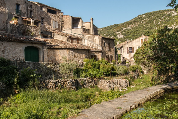 Altes Dorf mit einem kleinen Bach, Gras, Pflanzen, Bäumen, einer Steinmauer, Gebäuden mit Fenstern und Türen, Geländern, Hügeln und einem klaren blauen Himmel.