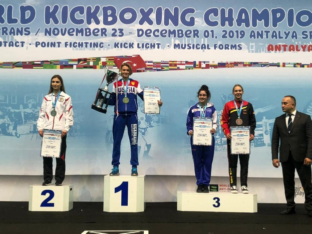 Fünf Personen auf einem Podium mit Trophäen und Urkunden vor dem Hintergrund einer "World Kickboxing Championship Antalya 2019"-Plakate.