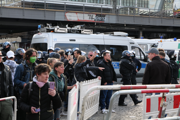 Eine Gruppe von Menschen steht vor einer Reihe von Polizeiwagen, einige tragen Helme und halten Mobiltelefone, mit Absperrungen im Vordergrund und einer Brücke mit Geländern und Gebäuden im Hintergrund, in Berlin, Deutschland.