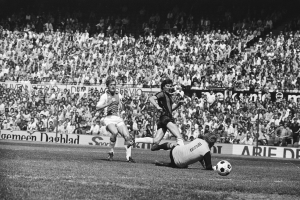 Schwarzes und weißes Foto von Männern, die auf einem Feld Fußball spielen, mit Zuschauern im Hintergrund und einem Banner "Bundesliga 1978-79 Ajax 1-1 Hannover 96".