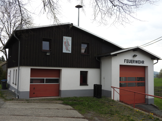 Feuerwache mit rotem und weißem Gebäude, Straße, Gras, gelben Blumen, schwarzen Objekten, Geländer, Pfahl, Drähte, Bäumen und Himmel sichtbar.