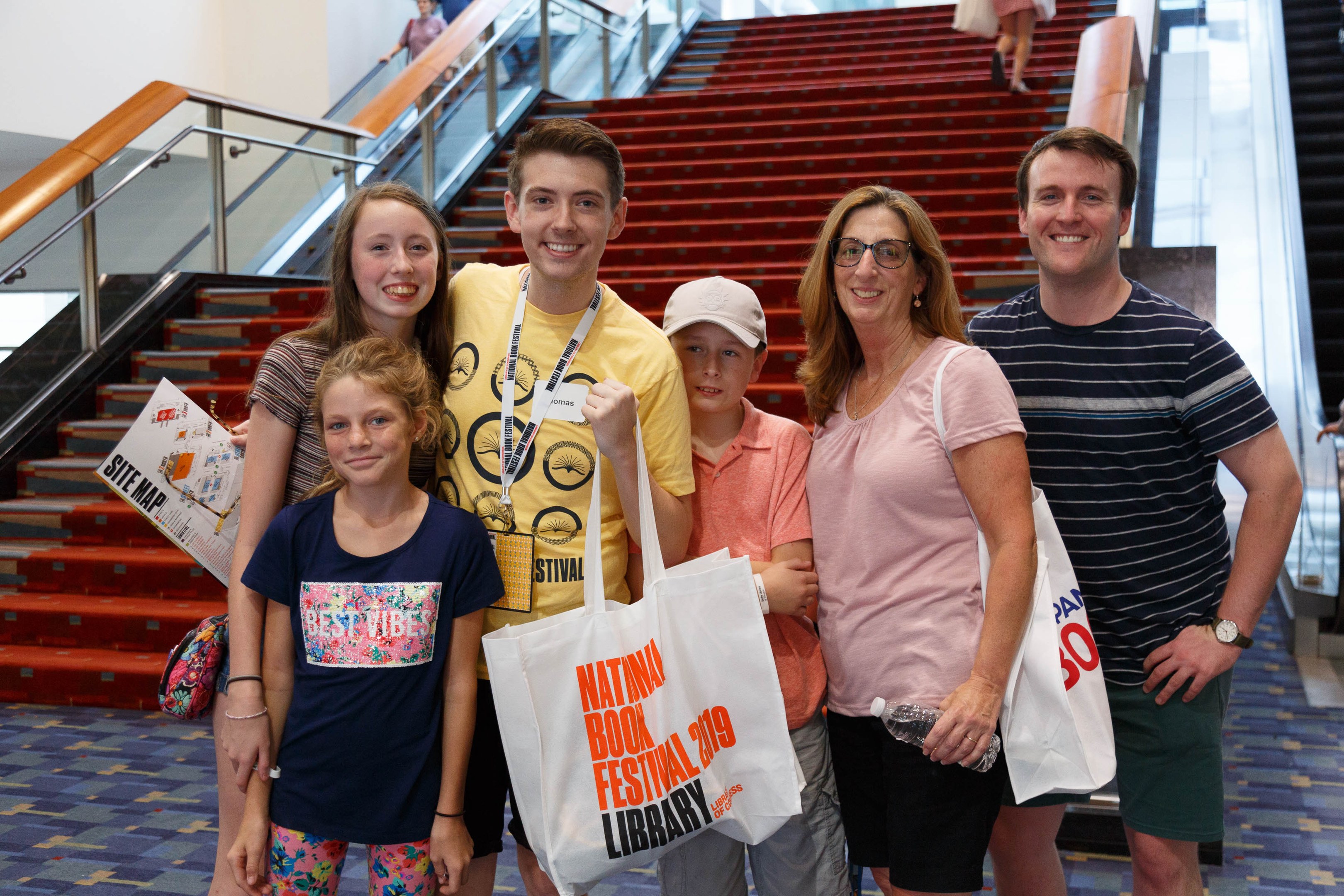 Eine Gruppe von Menschen, die vor einer Rolltreppe an der Nationalen Buchmesse 2019 lächeln und Taschen halten, mit Treppen, Geländern und einer Wand im Hintergrund.