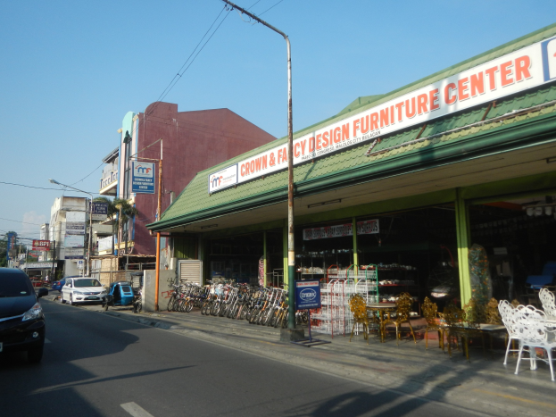 Eine belebte Stadtstraße mit fahrenden Fahrzeugen, parkenden Fahrrädern, Gebäuden mit Schildern, Laternen und Stromleitungen, mit einem Möbelgeschäft im Vordergrund und Himmel im Hintergrund.