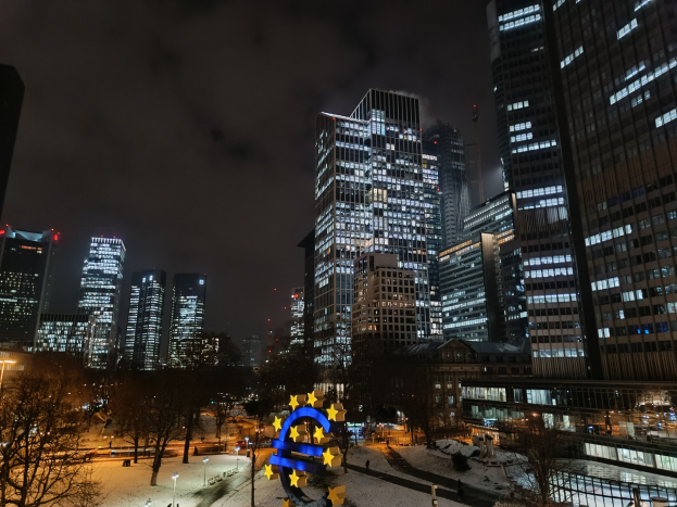 Europäische Zentralbank-Hauptquartier in Frankfurt, Deutschland bei Nacht, mit beleuchteten Gebäuden, Bäumen, Straßenlaternen, Lichtern und Fußgängern unter einem bewölkten Himmel.