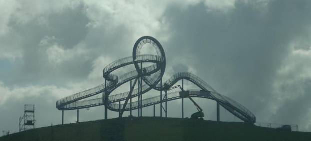 Eine Achterbahn auf dem Höhepunkt eines Hügels mit einem bewölkten Himmel im Hintergrund, umgeben von Stützpfählen und anderen Strukturen.