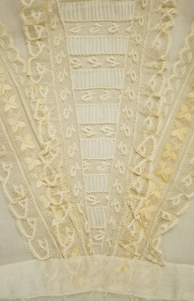 Nahaufnahme eines weißen Kleides mit zarter, detallierter Spitzenverzierung mit verschiedenen Formen und Mustern auf einem leichten, luftigen Stoff.
