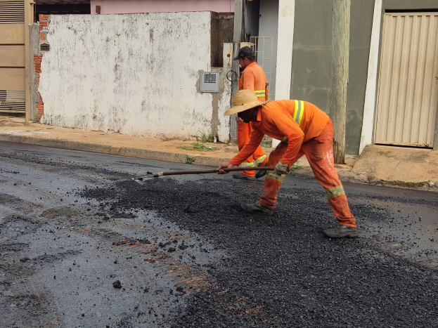 Zwei Männer in orangefarbenen Overalls und Hüten arbeiten auf einer Straße, wobei einer ein Objekt hält, vor Gebäuden mit Läden.