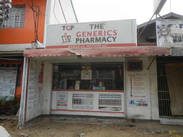 Außenansicht der Generics Pharmacy-Filiale mit umliegenden städtischen Elementen unter einem klaren blauen Himmel.