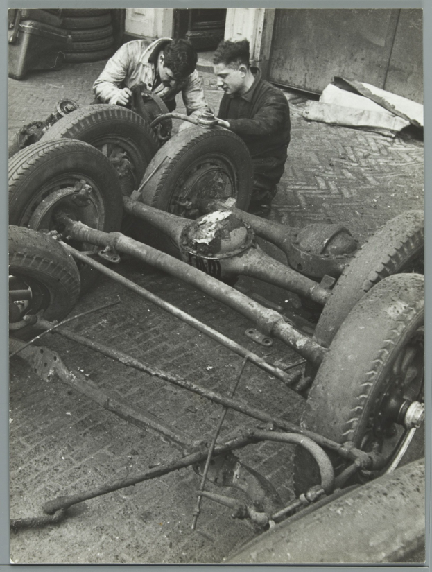 Zwei Männer in einer schwarzen und weißen Garage reparieren ein Auto und halten Werkzeuge, mit Reifen und anderen Gegenständen drumherum.