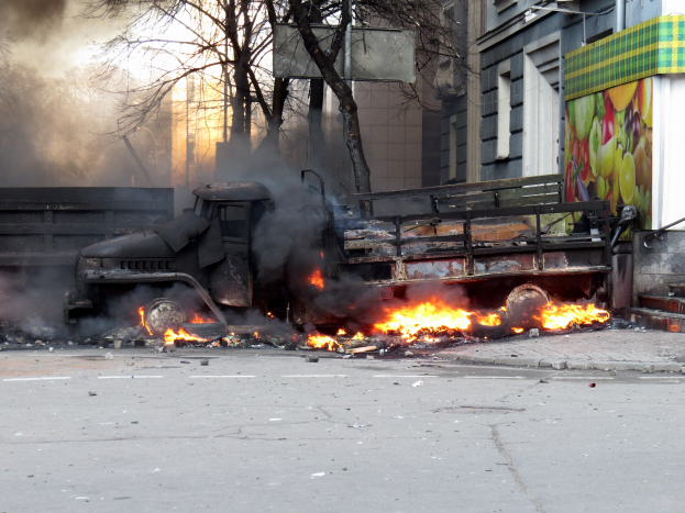 Ein großer Lastwagen brennt am Straßenrand mit dicken Rauchwolken, im Hintergrund sind Bäume und Gebäude unter einem klaren Himmel zu sehen.