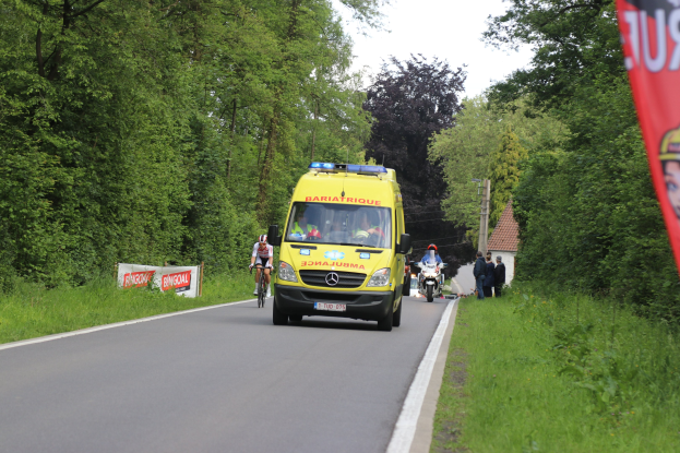 Ambulanz fährt auf einer Straße mit Radfahrern daneben, Gras und Bäume auf beiden Seiten, Häuser und Pfosten im Hintergrund unter einem klaren blauen Himmel.