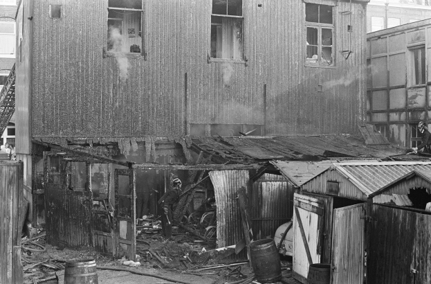 Ein Schwarz-Weiß-Foto eines durch Feuer zerstörten Gebäudes mit verstreuten Trümmern, darunter Fässer und hölzerne Gegenstände, sowie einigen Menschen in der Nähe, mit anderen Gebäuden, einer Leiter und einem Pfahl im Hintergrund.