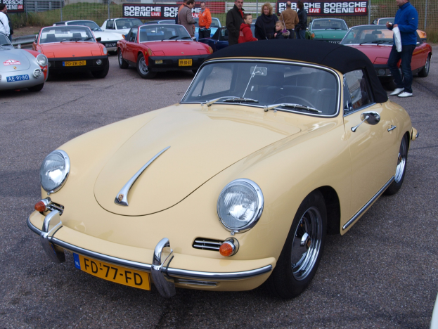 Gelber Porsche 356 Speedster auf einem Parkplatz mit Menschen in der Nähe, umgeben von anderen Autos, einem Zaun, Bannern, Bäumen, Gebäuden und einem klaren blauen Himmel.