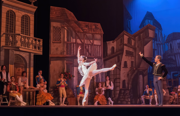 Zwei Tänzer performen auf der Bühne der Royal Opera House in London, mit einem Publikum im Vordergrund, das auf Stühlen sitzt und steht, und einer Kulisse aus Gebäudeillustrationen.
