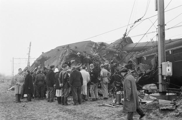 Schwarz-weißes Bild von Menschen mit Mützen in der Nähe eines Zuges, der gegen einen Pfahl gekracht ist, mit Strommasten, Drähten, Bäumen und einem klaren Himmel im Hintergrund.