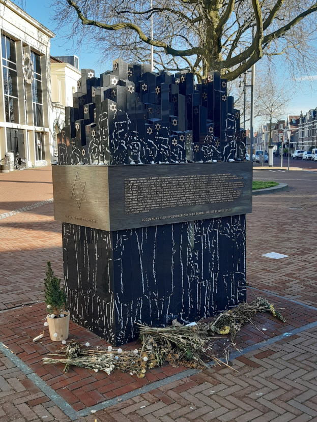 Denkmal in einem Stadtplatz, das Holocaust-Opfern gewidmet ist, mit einem Baum davor, einem Topfpflanzen auf dem Boden, verstreuten trockenen Blättern und Gebäuden, Fahrzeugen, Masten und Bäumen im Hintergrund.