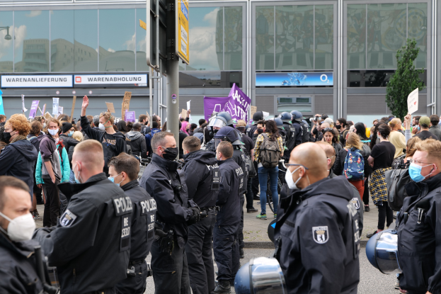 Eine große Gruppe von Menschen steht vor einem Gebäude, einige halten Schilder und tragen Helme, mit einer Stange und einem Schild im Vordergrund und einem Baum im Hintergrund, was auf eine Demonstration hindeutet.