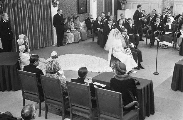 Schwarzes und weißes Foto einer Hochzeitszeremonie mit sitzenden und stehenden Menschen, einem Mann mit Mikrofon und einer Frau mit Blumenstrauß, mit einem Podium, Blumengestecken, einem Vorhang, einem gerahmten Foto an der Wand und einer Tür im Hintergrund.