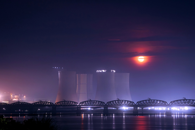 Atomkraftwerk bei Nacht mit Brücke über Wasser, beleuchtet von Gebäuden und Mondlicht, umgeben von Bäumen.