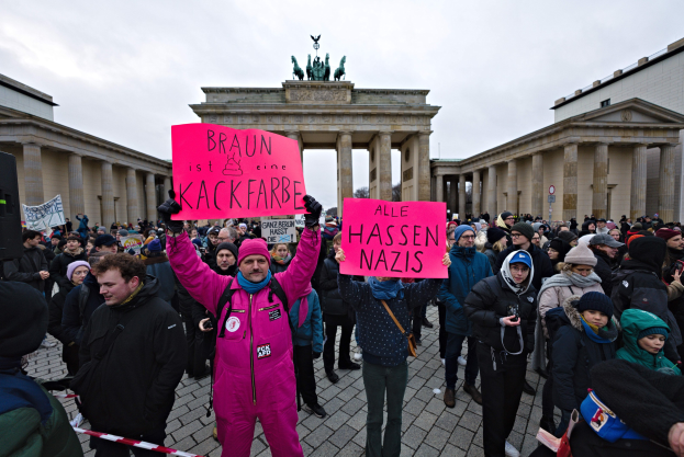 Gruppe von Menschen protestiert vor dem Brandenburger Tor, hält Schilder gegen die Anti-Nazis-Bewegung der deutschen Regierung, mit Gebäuden, Säulen und Statuen im Hintergrund unter einem klaren blauen Himmel.