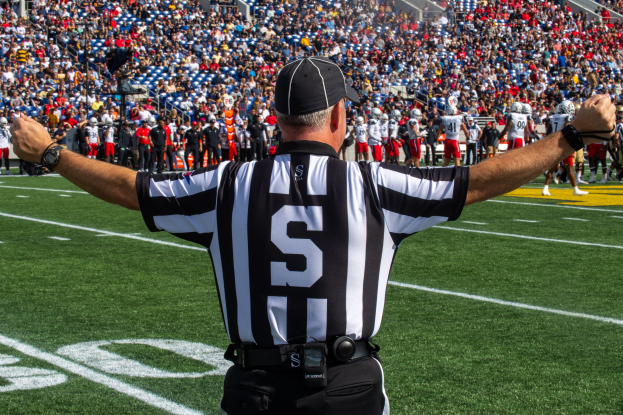 Schiedsrichter steht auf Fußballfeld mit ausgestreckten Armen, umgeben von Menschen auf dem Boden und einer sitzenden Menge im Hintergrund.