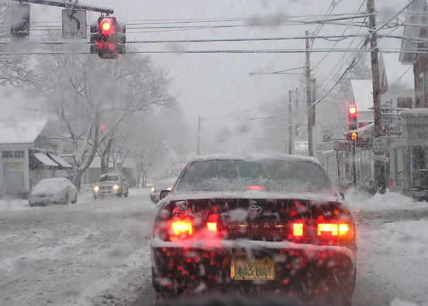 Wenige Fahrzeuge auf einer schneebedeckten Straße, die von schneebedeckten Bäumen und Häusern gesäumt ist.
