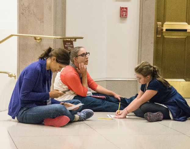 Drei Frauen sitzen auf dem Boden vor einer Tür, umgeben von Büchern und Schreibmaterialien, in Diskussion.