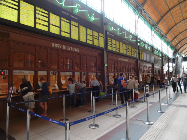 Menschen an einem Bahnhofsschalter mit übersichtsleuchten, Informationsanzeigen und Treppen mit Geländern.