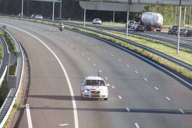 Ein Auto auf einer Autobahn mit einem stationären Polizeiwagen am Straßenrand, andere Fahrzeuge, Geländer, Pfosten, Schilder und Bäume im Hintergrund.
