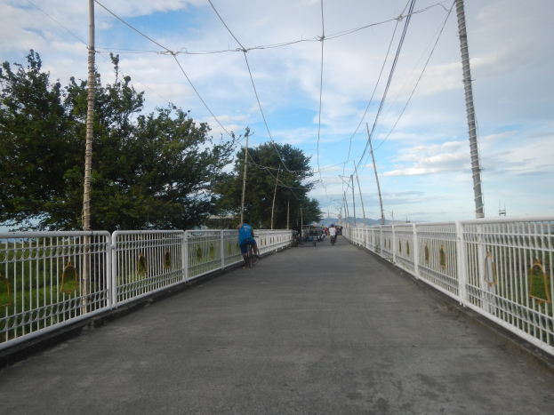 Eine Fußgängerbrücke mit Fahrradfahrern, Geländern, Strommasten, Bäumen und einem bewölkten Himmel im Hintergrund.