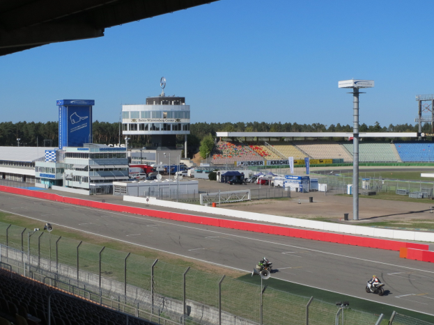 Eine Gruppe von Menschen auf Motorrädern auf einer Rennstrecke umgeben von einem Zaun, Fahrzeugen, einem Gebäude, Schildern, Stadion und Bäumen unter einem bewölkten Himmel.