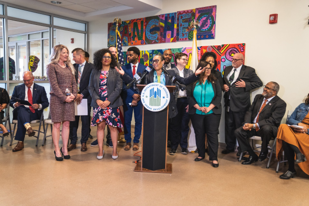 Eine Gruppe von Menschen steht vor einem Podium mit einem Mikrofon und einer Tafel mit dem Text "Sancho Stadtverordneter tritt dem Stadtrat bei", während andere auf Stühlen sitzen oder in der Nähe stehen. Flaggen, eine Tafel, ein Feuerlöscher und Deckenleuchten sind an der Rückwand zu sehen.