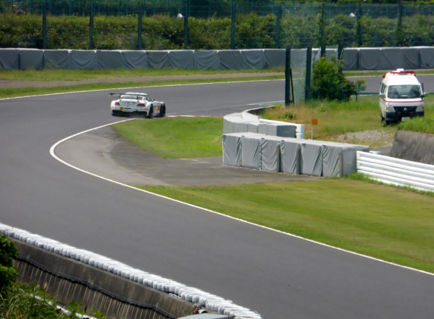 Ein Rennauto auf einer Rennstrecke mit einem Polizeiwagen im Hintergrund, umgeben von Gras, Pflanzen, Bäumen und Absperrungen, mit einem Fahrzeug auf der rechten Seite und Text unten.