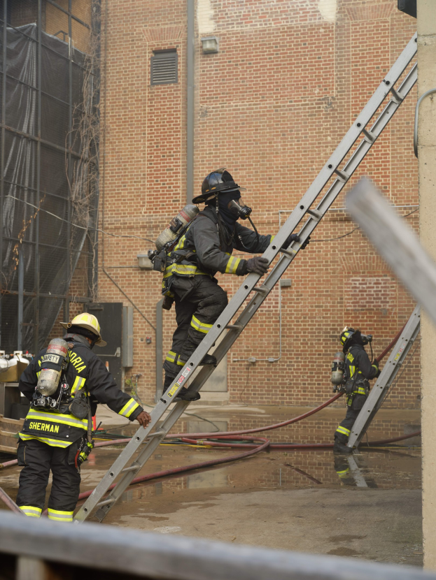 Feuerwehrleute in Helmen und Ausrüstung, die eine Leiter hochklettern, vor einem Backsteingebäude mit Rohren auf dem Boden und einer Metallstange am unteren Ende, mit einem weiteren Gebäude und einem Netz im Hintergrund.