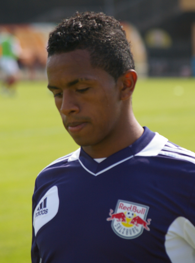 Ein junger Mann in einem blauen T-Shirt steht auf einem Fußballfeld, der als Flügelspieler mit kurzen dunklen Haaren und einem entschlossenen Gesichtsausdruck gegen einen unscharfen Hintergrund erscheint.