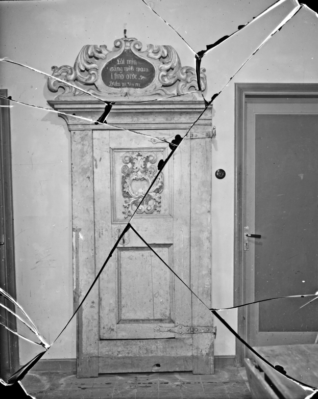 Schwarz-weiß-Foto einer zentral platzierten gebrochenen Tür mit verstreuten Glasscherben in einem Raum mit einer sichtbaren Wand im Hintergrund.