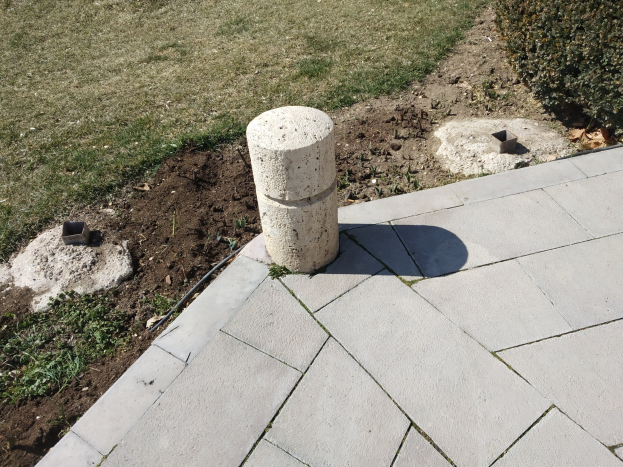 Betonpoller auf einem Ziegelweg mit grünem Gras und Pflanzen drumherum.