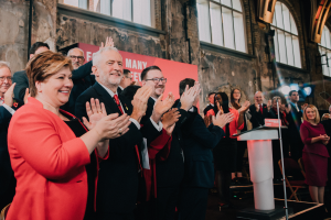 Eine Gruppe von Menschen, wahrscheinlich Liberale, klatscht feiernd vor einer Menge, mit einem Podium, Mikrofon und Texttafel rechts, Stühlen, einer Fahne, einer Wand mit Fenstern und Lichtern im Hintergrund.