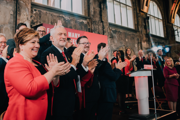 Eine Gruppe von Menschen, wahrscheinlich Liberale, klatscht feiernd vor einer Menge, mit einem Podium, Mikrofon und Texttafel rechts, Stühlen, einer Fahne, einer Wand mit Fenstern und Lichtern im Hintergrund.