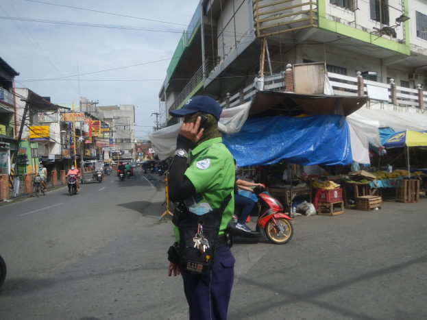 Polizist in grüner und blauer Uniform mit blauer Mütze, der auf einer Straße steht und auf einem Handy spricht, mit Fahrzeugen, Marktständen, einem Gebäude und bewölktem Himmel im Hintergrund.