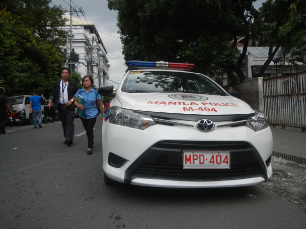 Ein Polizeiwagen am Straßenrand mit einem sichtbaren Nummernschild, einige Menschen auf dem Gehweg, ein Geländer und eine Wand auf der rechten Seite und Bäume, Gebäude, Pfosten, Drähte und Himmel im Hintergrund.